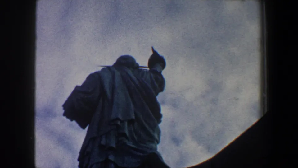 Low-angle 1960 New York City 8mm view of the Statue of Liberty, with the torch and robe visible against clouds.