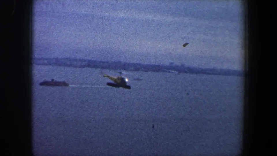 1960 New York City harbor view showing a helicopter over water, a small vessel, and distant shoreline in grainy 8mm color.