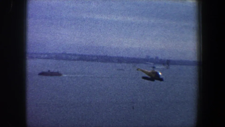 1960 New York City 8mm frame of a helicopter flying over harbor water, with a hazy shoreline visible beyond.
