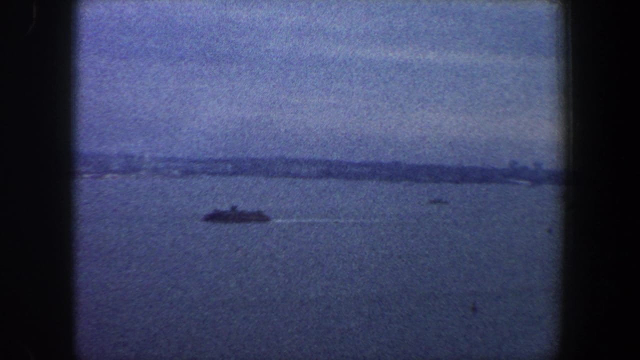 1960 New York City harbor view with a distant ship crossing open water under a hazy sky in blue-tinted 8mm footage.
