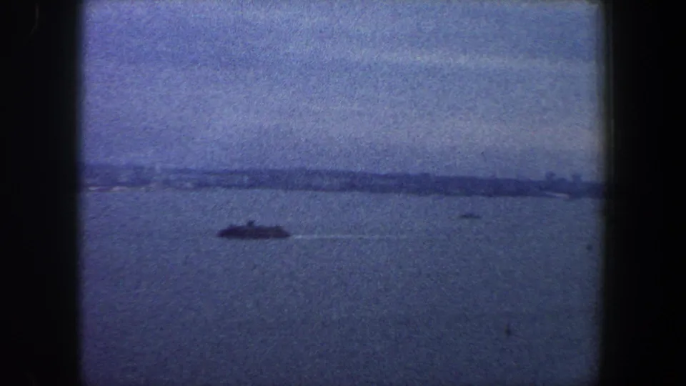 1960 New York City harbor view with a distant ship crossing open water under a hazy sky in blue-tinted 8mm footage.