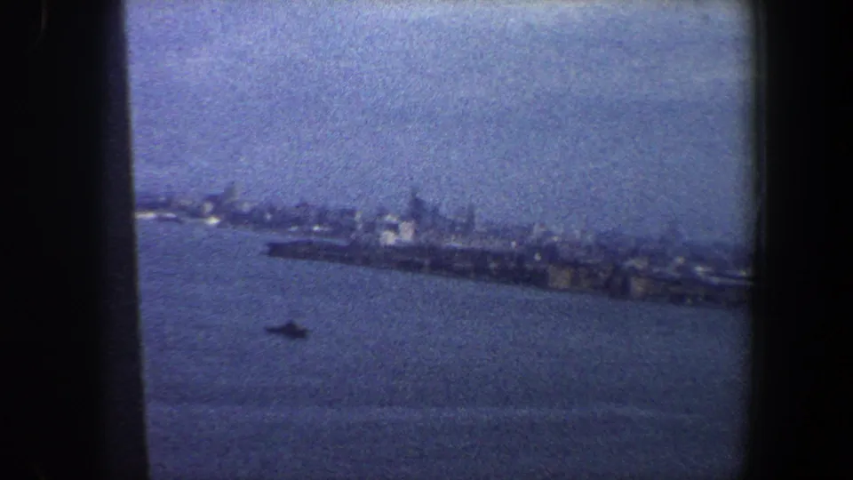 Tilted 8mm view of the New York City waterfront in 1960, with buildings along the shore and a small boat on the water.