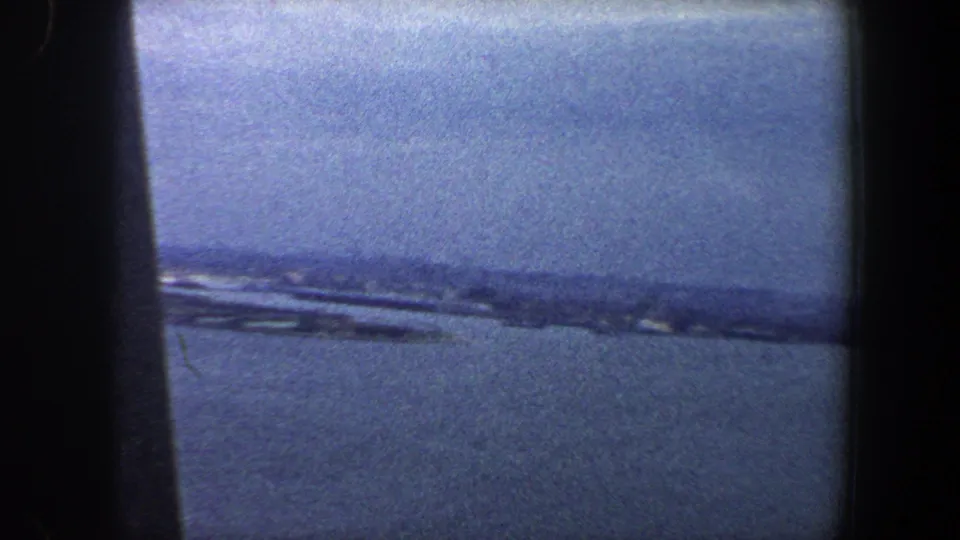 1960 8mm New York City coastal view with blue water, hazy sky, and a distant shoreline framed by dark gate edges.