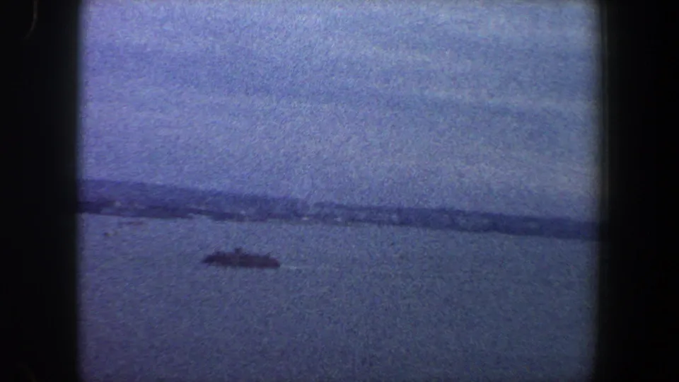 1960 8mm New York City waterfront view with a dark boat low in frame and a faint shoreline under a pale sky.