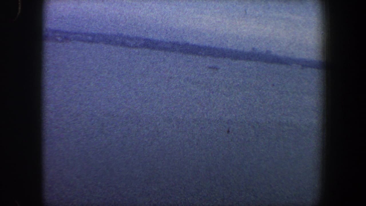 1960s New York City 8mm frame showing open water, a faint shoreline, blue sky, grain, and dark scan borders.