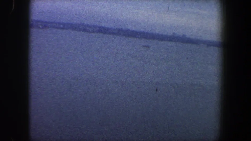 1960s New York City 8mm frame showing open water, a faint shoreline, blue sky, grain, and dark scan borders.