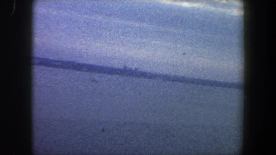 1960s New York City 8mm frame with water, a low distant shoreline, pale sky, and heavy film grain.