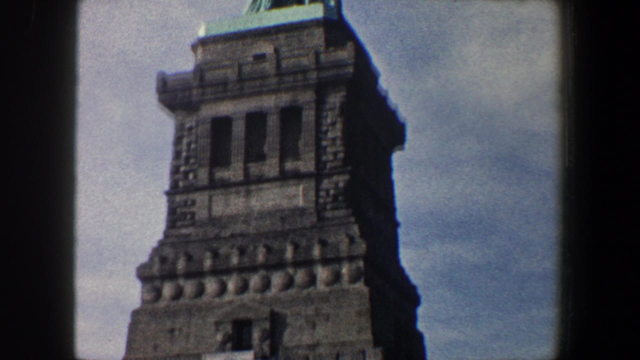 1960 8mm home-movie frame from New York City showing the Statue of Liberty pedestal and lower portion of the statue against a cloudy sky.