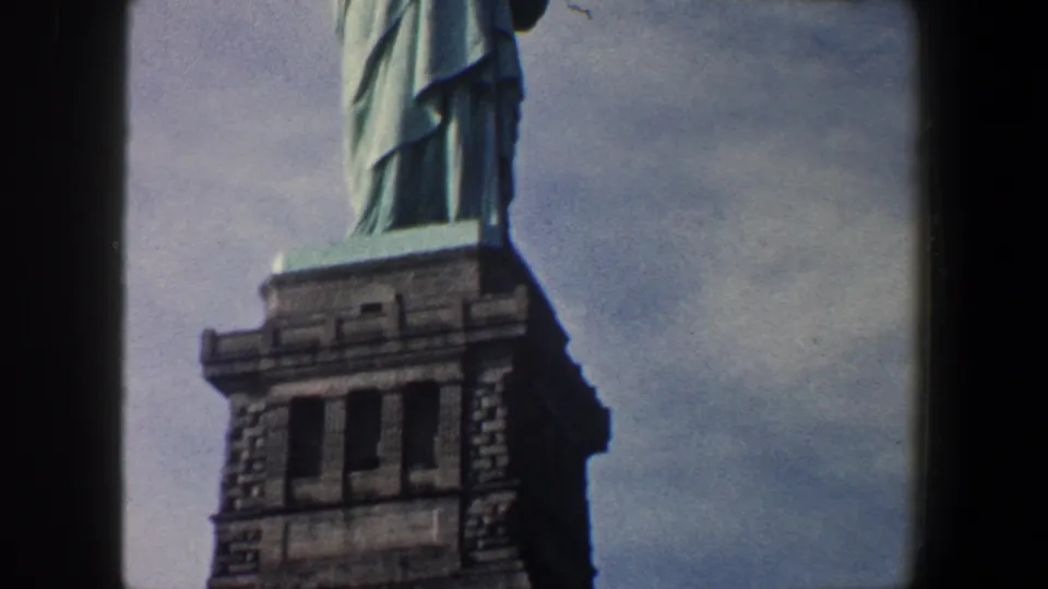 1960 New York City home-movie frame showing the upper stone pedestal and lower section of the Statue of Liberty during an upward tilt.