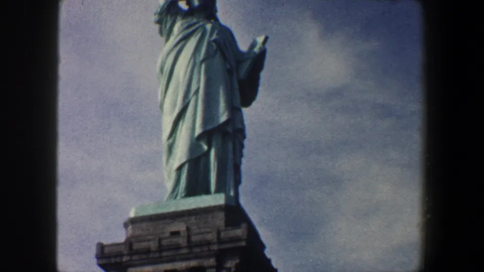 1960 8mm frame of the Statue of Liberty in New York City, with the robed figure partly framed above the pedestal against a muted cloudy sky.