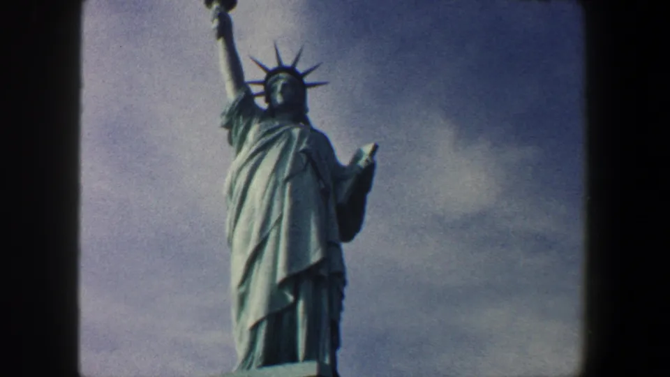 1960 New York City 8mm home-movie frame showing the Statue of Liberty from pedestal to crown, with the raised torch arm and tablet visible against a cloudy sky.