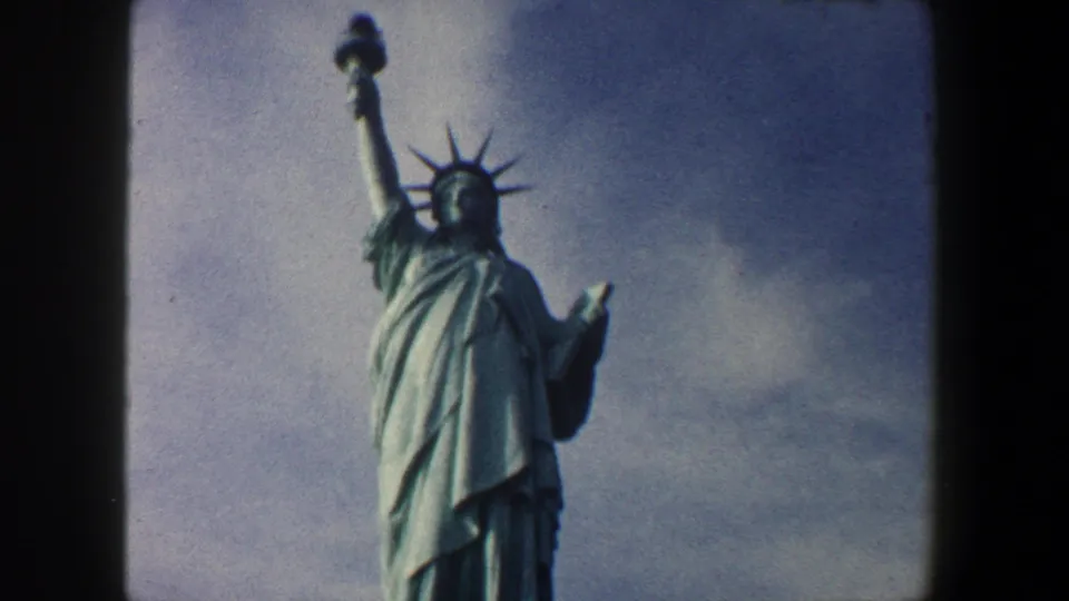 1960 8mm New York City footage showing the Statue of Liberty from below, with torch, crown, robe, and cloudy sky visible.
