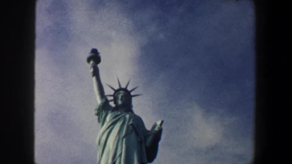 1960 8mm footage frames the Statue of Liberty from below, with the torch, crown, and tablet visible beneath a textured cloudy sky.