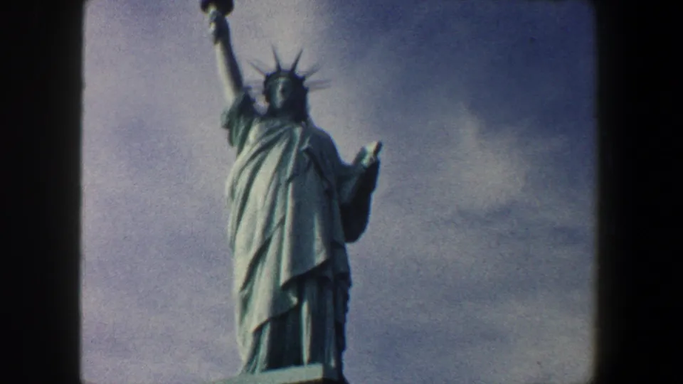 The Statue of Liberty is shown in 1960 New York City home-movie footage from a low angle, with robe folds, torch, crown, and pedestal edge visible.