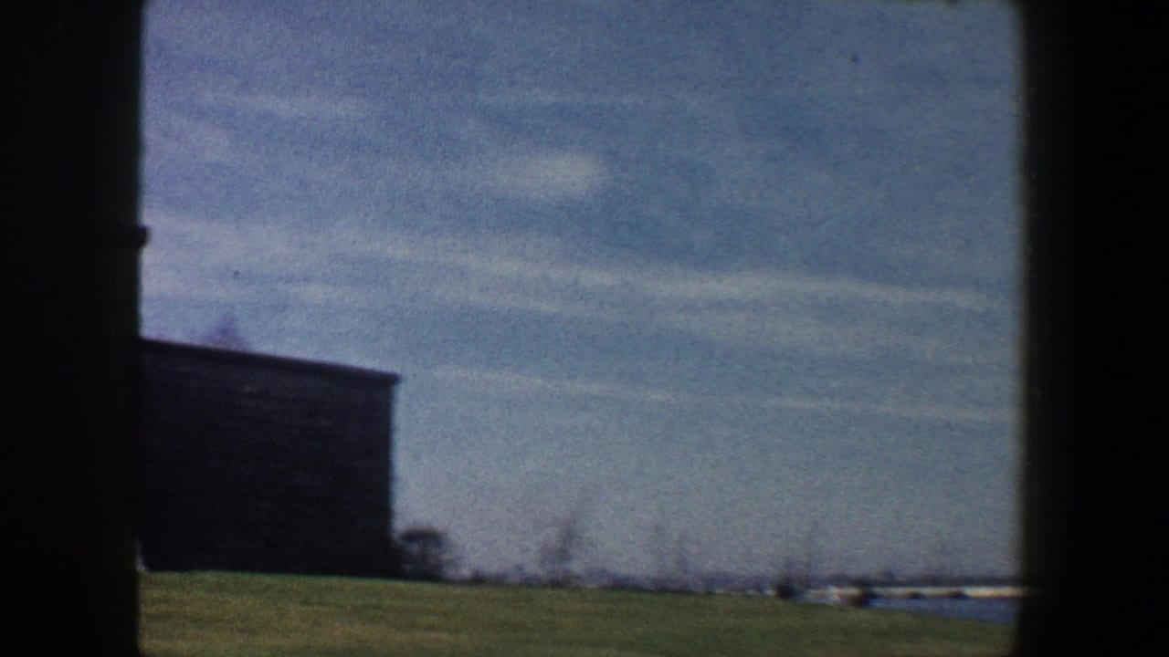 Dark building beside grass with water and distant shoreline under a blue sky.