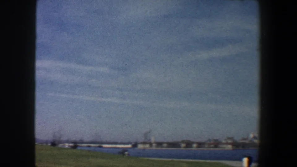 1960 New York City harbor-side view across grass and water toward low distant buildings and skyline forms.