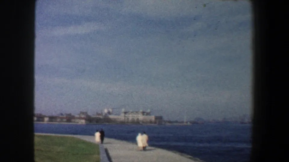 1960 New York City waterfront promenade with two distant pedestrians on a paved path, grass, harbor water, and shoreline buildings.