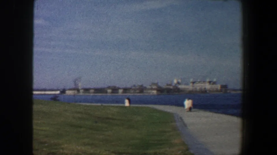 1960 New York City waterfront promenade view with a curving path, grassy bank, open water, and low buildings on the far shore.