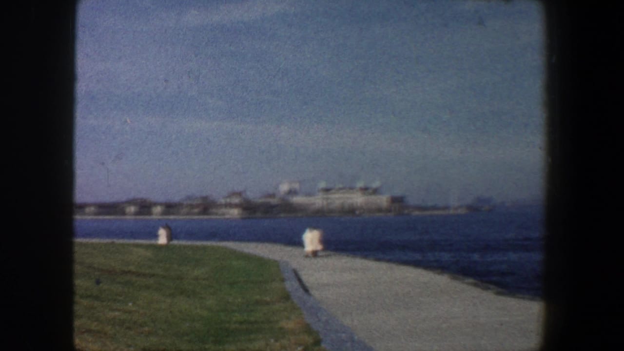 1960 New York City waterfront view with grass, a paved path, harbor water, distant buildings, and two small figures walking away.