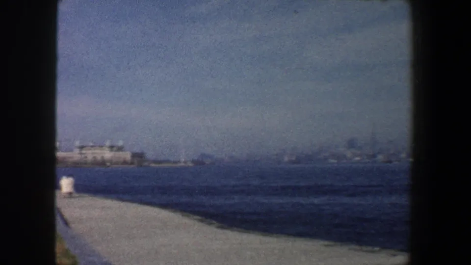 1960 New York City harbor view from a waterfront path, with distant low buildings and hazy skyline forms across the water.