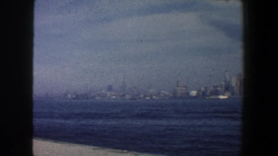 Hazy 1960 New York City skyline seen across blue harbor water, with a pale cloudy sky and soft 8mm film texture.