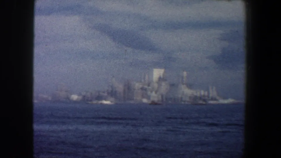 1960 New York City skyline across the harbor, with tall buildings softened by haze and visible grain from the 8mm film scan.