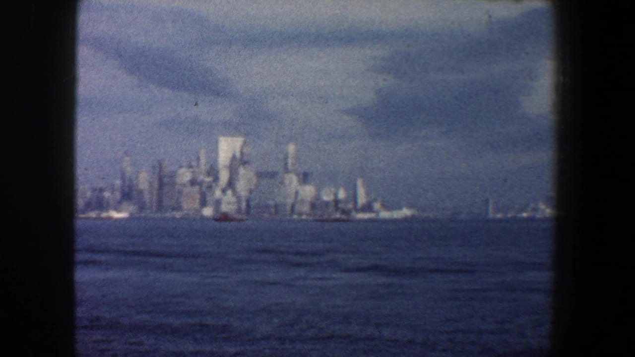 Distant New York City skyline across blue water with small boats and hazy clouds.