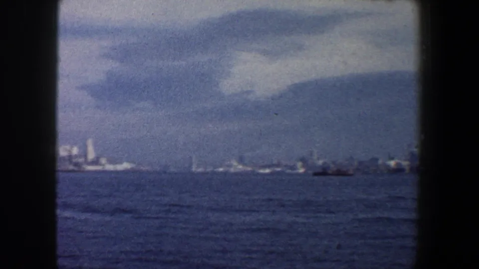 1960 New York City waterfront view with soft distant buildings, open blue water, and a small boat near the right horizon.