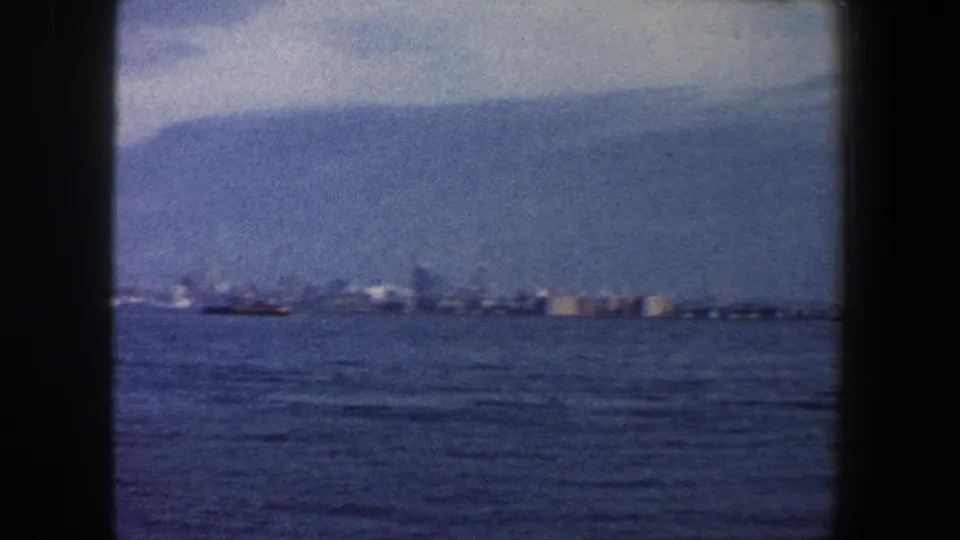1960 waterfront home-movie frame showing a low New York shoreline across blue water beneath a hazy, clouded sky.