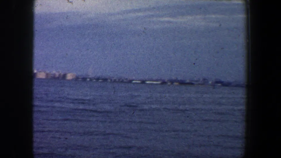 1960 8mm frame of a distant New York waterfront line across deep blue water, softened by haze and film grain.