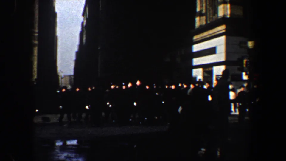 1960 New York City home-movie frame showing pedestrians silhouetted on a wet nighttime street beside lit building facades.