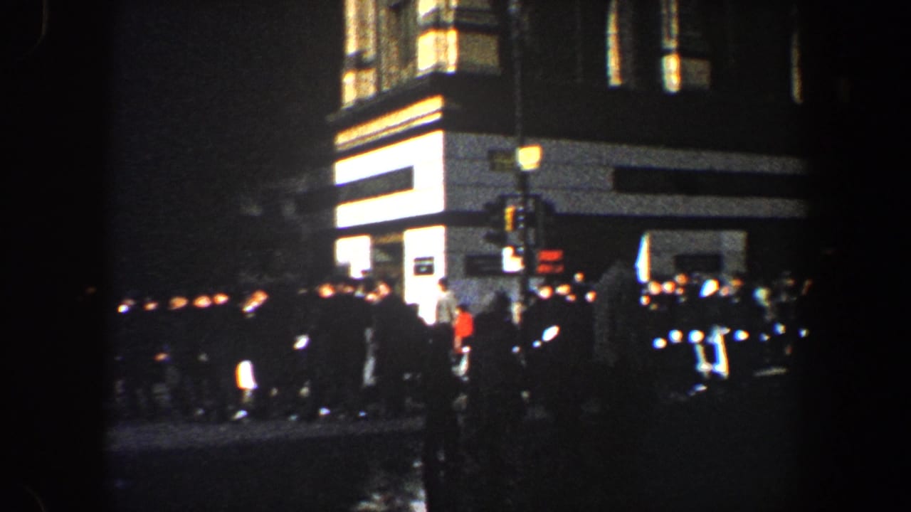 Dense nighttime crowd beside a bright corner building and wet street.