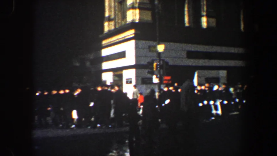 1960 New York City street crowd at night, with wet pavement, lit windows, traffic signals, and dark vehicles near a corner building.