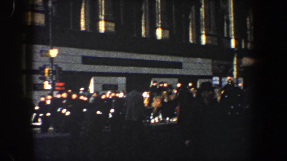 1960 New York City frame with pedestrians, dark cars, wet pavement, and a bright building entrance in low-light 8mm footage.