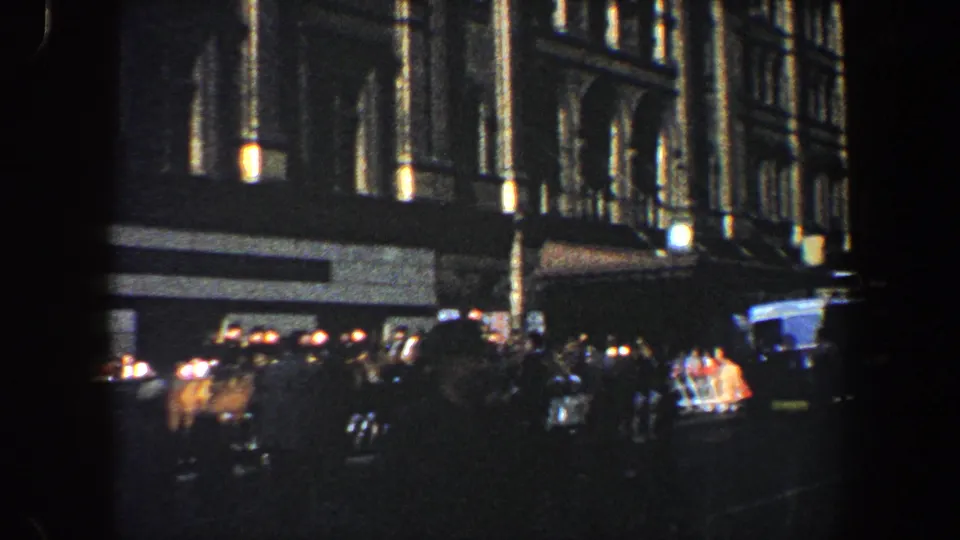 1960 New York City nighttime street scene with pedestrians, cars, wet pavement, and illuminated mid-century building windows.