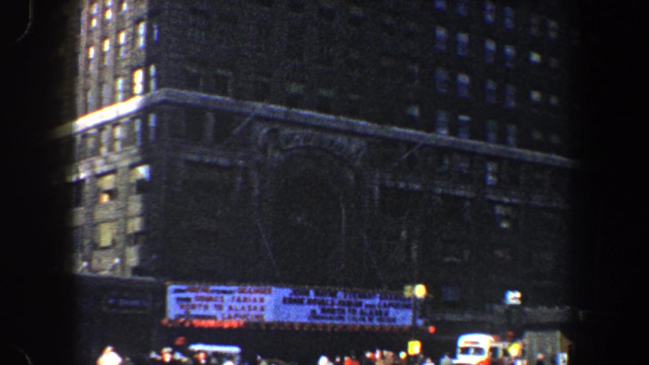 Dark city block with tall buildings, a lit marquee, pedestrians, and street traffic.