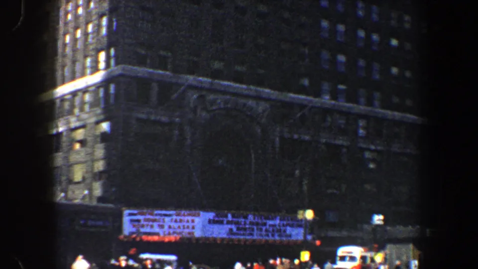 1960 New York City street view with high-rise facades, a theater-style marquee, pedestrians, and vehicles seen from a handheld 8mm frame.