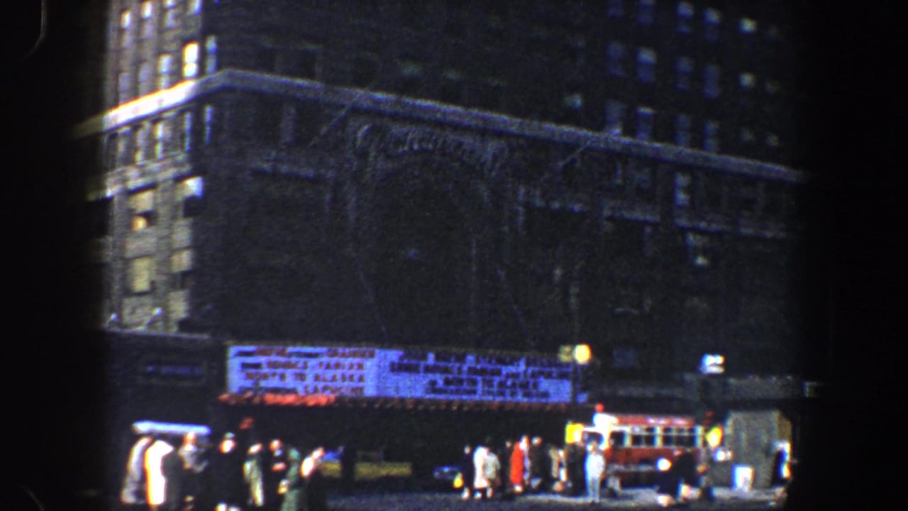 Busy street scene beneath high buildings with a marquee, pedestrians, cars, and a red bus.