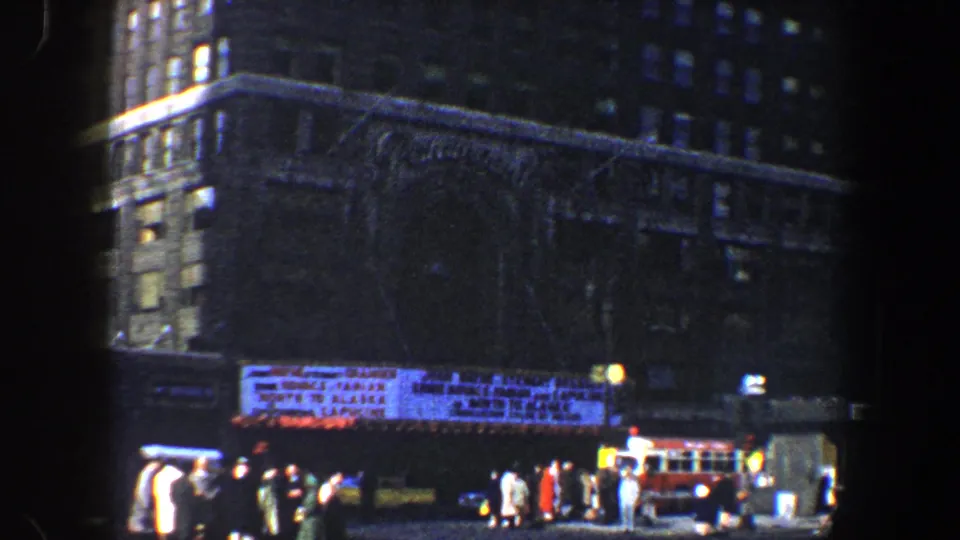 1960 New York City street scene showing a crowded sidewalk, theater-style marquee, high-rise buildings, cars, and a red bus.