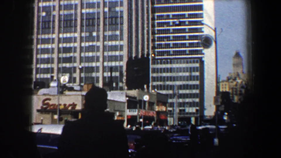 1960 New York City street view with modern high-rises, storefront signage, traffic, and a silhouetted pedestrian in the foreground.