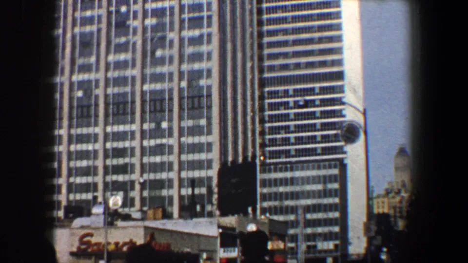 1960 New York City frame showing office towers, streetlights, partial storefront signage, and a dark pedestrian silhouette at street level.