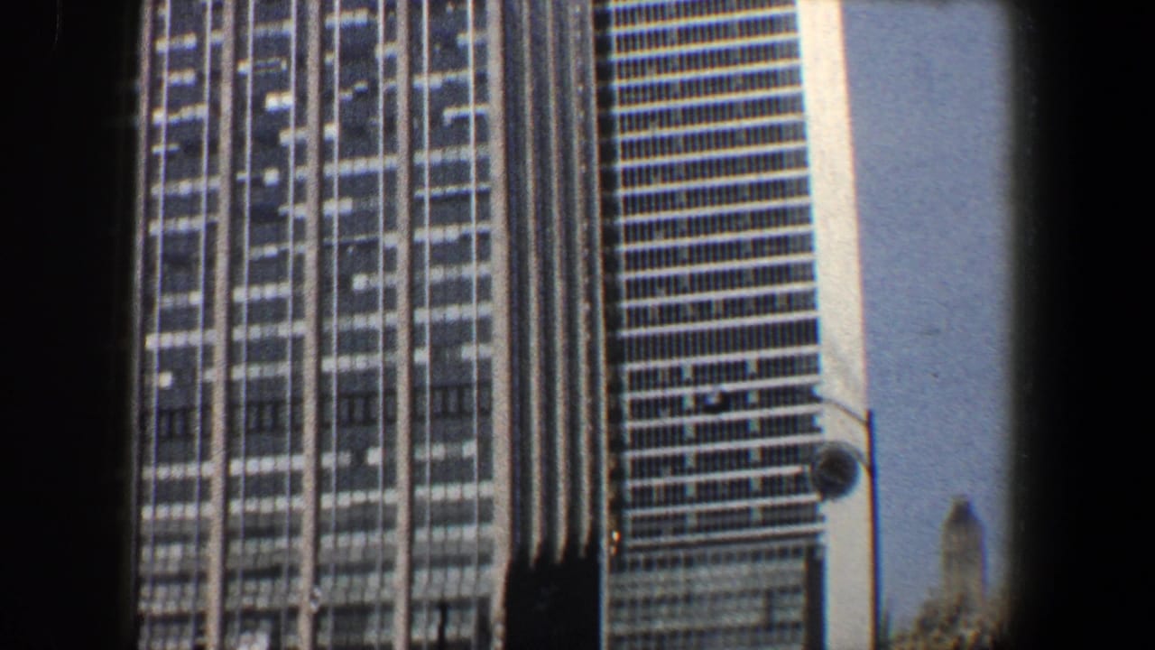 1960 New York City home-movie frame showing a tall glass-and-steel skyscraper facade and adjacent tower edge under bright sky.