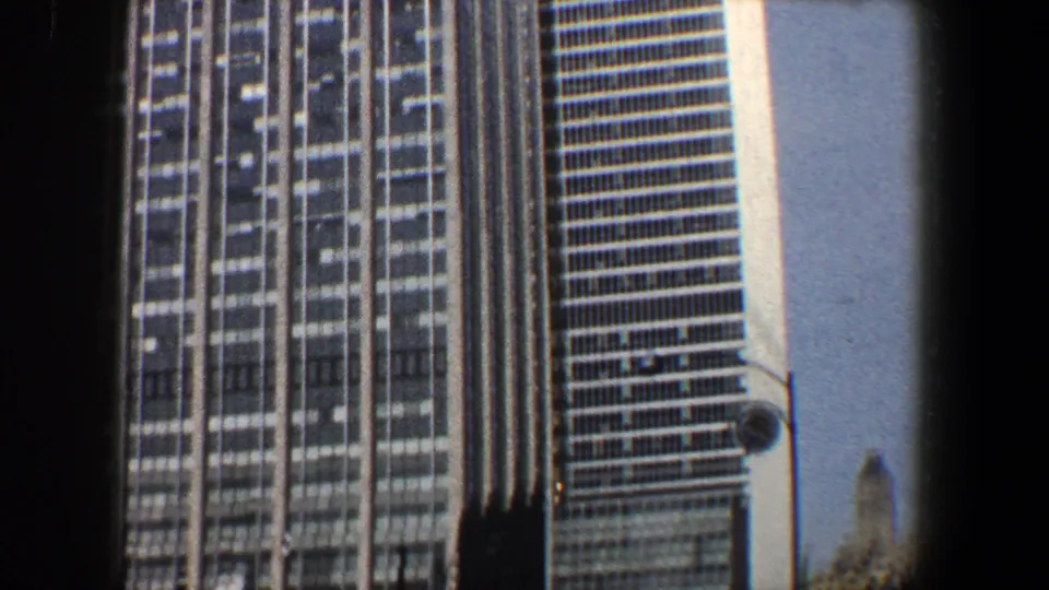 1960 New York City home-movie frame showing a tall glass-and-steel skyscraper facade and adjacent tower edge under bright sky.