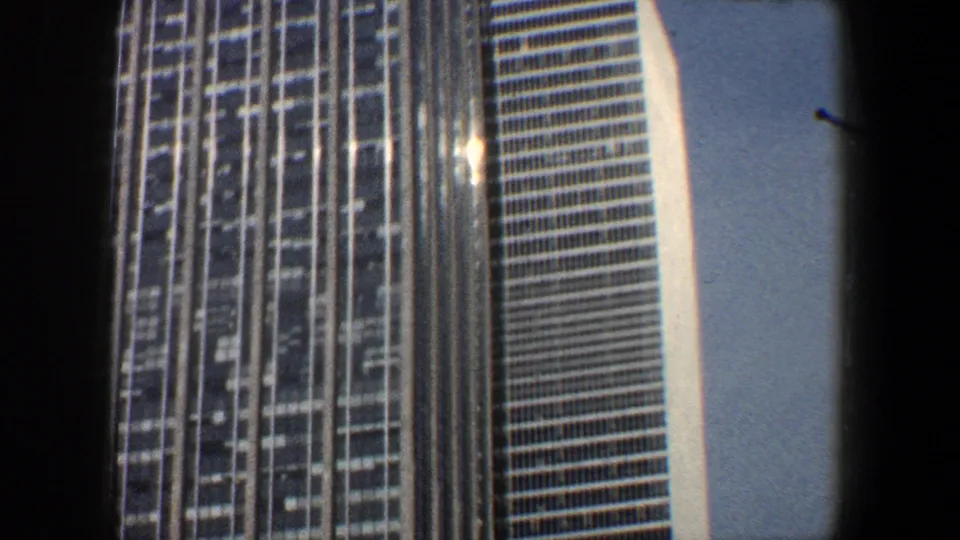 1960 New York City frame looking up between modern glass towers, with a streetlight and blue sky visible below the facades.