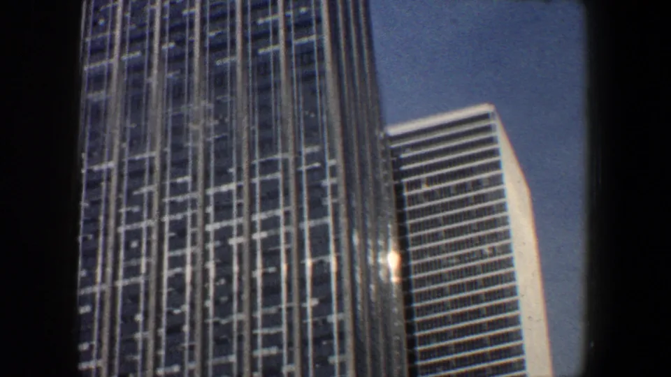1960 New York City home-movie frame showing two tall modern buildings from street level, with dense window grids and bright sky.
