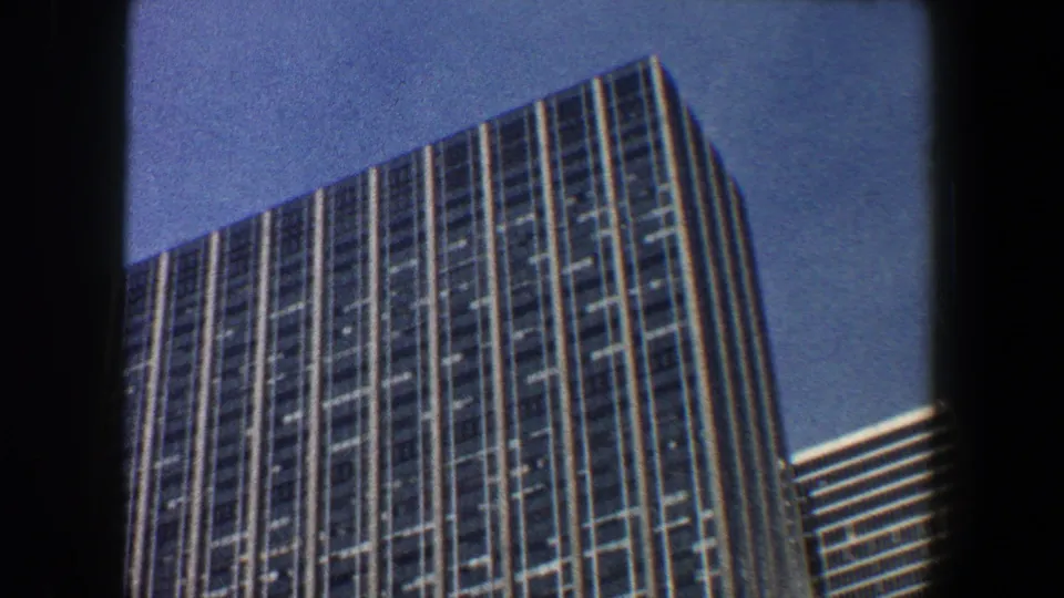 1960 New York City frame with a tilted upward view of a glass skyscraper crown and a neighboring striped high-rise under blue sky.