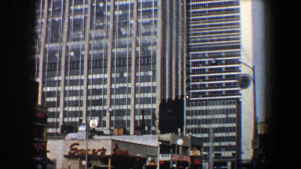 1960 New York City frame with a tall gridded building, street signals, neighboring tower, and partial storefront signage.