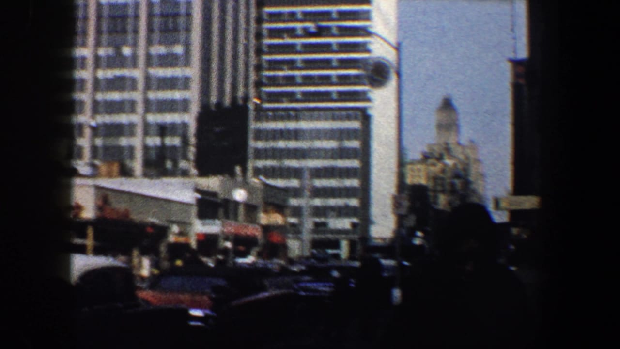 City street with tall buildings, parked cars, and dark pedestrian silhouettes in the foreground.