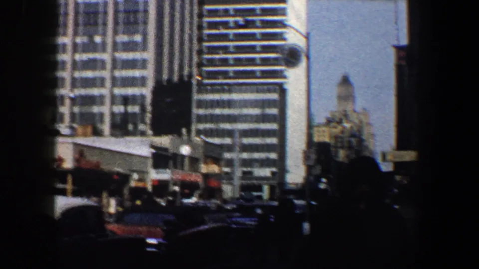 1960 New York City street view with mid-century high-rises, curbside cars, and pedestrians silhouetted by deep shadow.