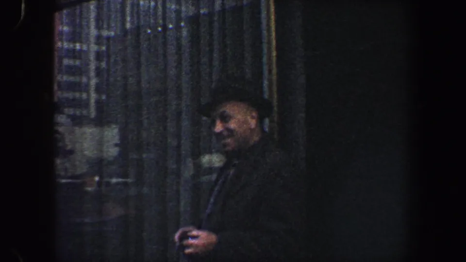 A smiling man in a dark hat and coat stands near a reflective city facade in 1960 New York street footage.